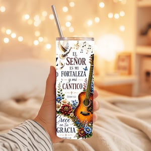 May include: A cylindrical tumbler with a metal straw featuring a graphic of an acoustic guitar, colorful flowers, a dove, and musical notes. The text reads: EL SEÑOR ES MI FORTALEZA y mi CÁNTICO, ÉXODO 15:2, Crece en Su GRACIA, 2 PEDRO 3:18.