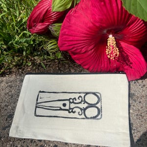 May include: A cream-colored zippered pouch with a black scissors design. The pouch is rectangular and lies on a concrete surface, with vibrant red hibiscus flowers in the background.