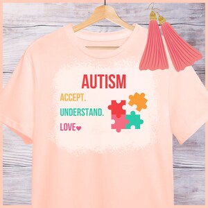 May include: Pink t-shirt with the words "AUTISM", "ACCEPT", "UNDERSTAND", and "LOVE" with a heart symbol. The shirt also features a colorful puzzle piece design.