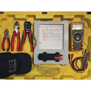 Op de afbeelding: Een gele plastic gereedschapskist met een selectie gereedschap, waaronder tangen, draadstrippers, een multimeter, veiligheidsbrillen en een zwarte baseballpet met de tekst "Phillips Industry". Een wit notitieblok met een handgeschreven takenlijst voor 3 december 2022 is ook zichtbaar.