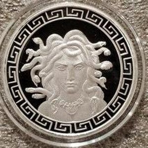 May include: A silver coin with a black background featuring a detailed image of Medusa, a Greek mythological figure with snakes for hair. The coin is surrounded by a Greek key border.