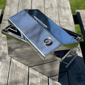 May include: A stainless steel smoker box with a lid and a built-in thermometer. The smoker box is sitting on a wooden surface.