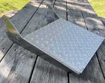 May include: A silver metal grilling grate with a diamond plate pattern. The grate has two metal brackets on the sides. The grate is sitting on a wooden surface.