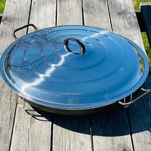 May include: A large, round, stainless steel pot with a lid. The pot has a black handle and the lid has a silver handle. The pot is sitting on a wooden surface.