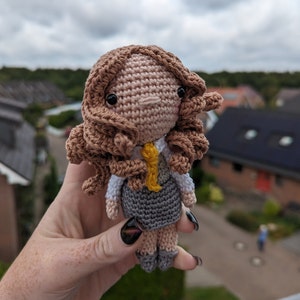Könnte beinhalten: Häkelpuppe mit braunen Haaren, einem weißen Hemd, einem grauen Rock und einer gelben Krawatte. Die Puppe hält eine gelbe Krawatte in der Hand.
