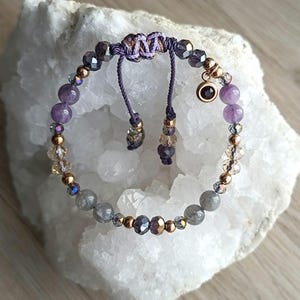 May include: A purple and grey beaded bracelet with a gold charm and a purple and gold cord closure.