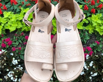 May include: A pair of white leather sandals with intricate floral designs. The sandals have a toe loop and an ankle strap with a buckle closure. The sandals are labeled "MADE IN MEXICO" and "MEX-4 USA-7".