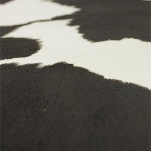 May include: Close-up of a black and white cowhide pattern. The design features irregular black shapes against a white background, creating a natural, textured appearance. The fabric has a soft, slightly fuzzy texture.