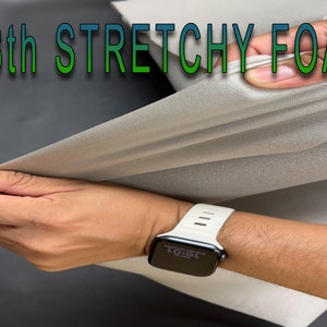 May include: A hand stretches a piece of 1/8th inch thick stretchy foam. The foam is light gray and has a slightly textured surface. The hand is wearing a white Apple Watch.