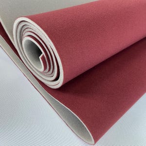 Puede incluir: Esterilla de yoga roja enrollada con una parte inferior gris claro. La esterilla está hecha de un material texturizado y está parcialmente desenrollada, revelando el interior gris claro. Los bordes son limpios y la apariencia general es suave.