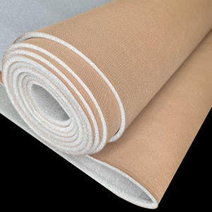 Puede incluir: Tela enrollada de color beige con bordes blancos. La tela está enrollada firmemente, revelando capas del material. Los bordes tienen una apariencia limpia y nítida. El fondo es negro.