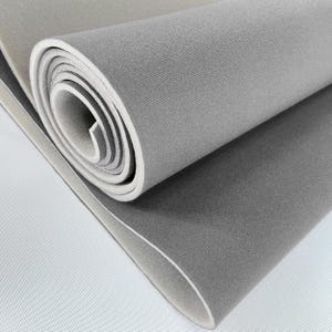 Puede incluir: Esterilla de espuma gris enrollada con bordes blancos. La esterilla está hecha de un material suave y texturizado. La sección enrollada revela el interior blanco, contrastando con el exterior gris. Probablemente utilizada para ejercicios o como superficie protectora.