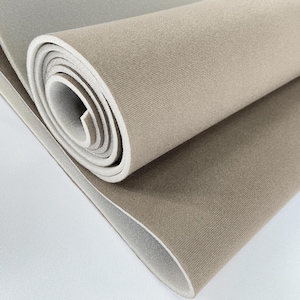 Puede incluir: Una sección enrollada de tela beige con interior blanco. La tela tiene una superficie texturizada y parece ser un material grueso, posiblemente para tapicería o manualidades. El rollo está parcialmente desenrollado, revelando la parte inferior blanca.