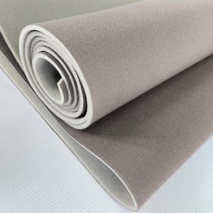 Puede incluir: Esterilla de yoga gris claro enrollada, con una parte inferior blanca. La esterilla está hecha de un material suave y texturizado y está parcialmente desenrollada, revelando el interior blanco. Los bordes son redondeados y la esterilla parece nueva.