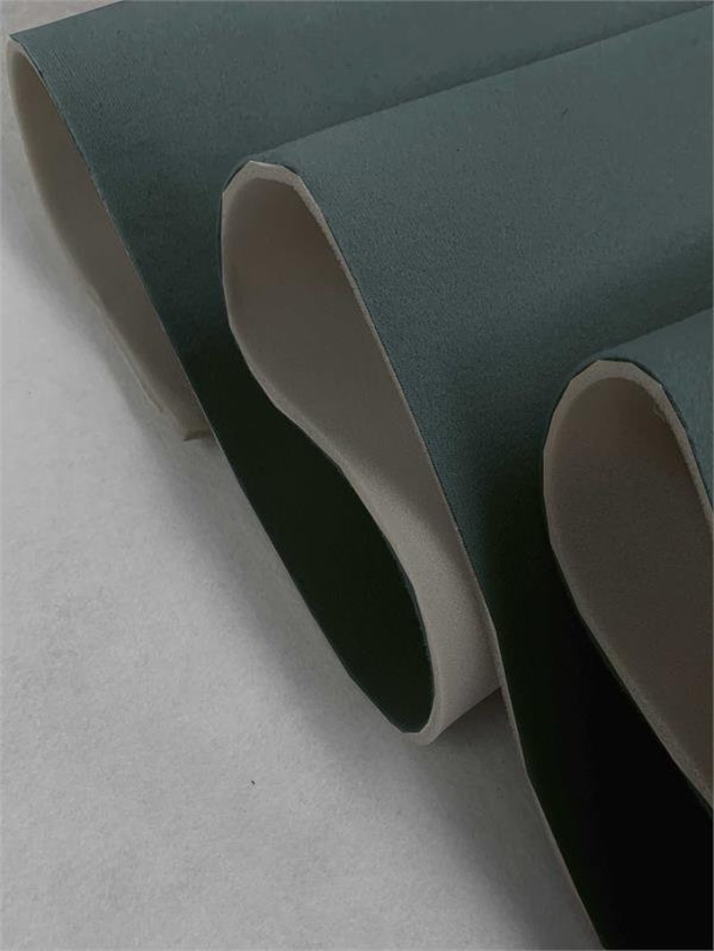 May include: Close-up of a dark teal fabric with a white backing. The fabric is folded over on itself, showing the white backing.