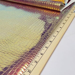 Op de afbeelding: Iriserende imitatieleer stof met een krokodillenleer textuur. Het materiaal is een mix van goud, paars en blauw. Een houten liniaal met inch markeringen staat op de voorgrond.
