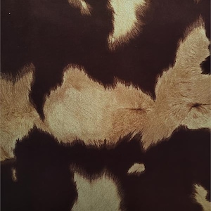 May include: Close-up of a cowhide pattern. The design features irregular patches of dark brown and tan, creating a natural, textured appearance. The fur has a soft, slightly raised texture.