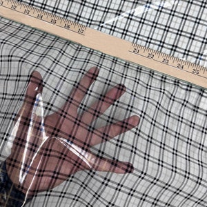 May include: Clear plastic sheet with a black and white plaid pattern. The sheet is draped over a white surface and a hand is visible through the plastic.