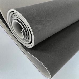 Puede incluir: Esterilla de yoga gris enrollada con un borde interior blanco. La esterilla está hecha de un material texturizado y está parcialmente desenrollada, revelando una superficie gris más clara. La imagen es un primer plano, destacando la textura y las variaciones de color.