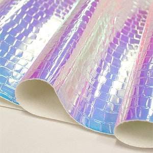 Op de afbeelding: Iridescent kunstleer met een krokodillenleertextuur. Het materiaal reflecteert licht en toont tinten paars, blauw en roze. De randen zijn opgerold en tonen een witte achterkant. Geschikt voor knutselprojecten.