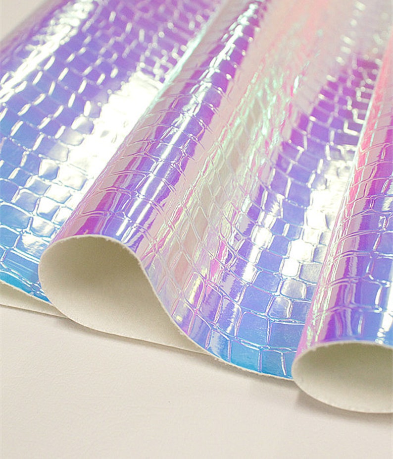 Op de afbeelding: Iridescent kunstleer met een krokodillenleertextuur. Het materiaal reflecteert licht en toont tinten paars, blauw en roze. De randen zijn opgerold en tonen een witte achterkant. Geschikt voor knutselprojecten.