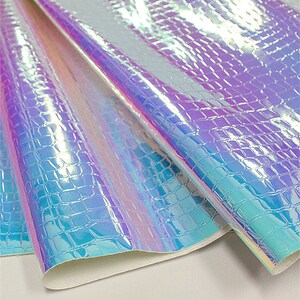 Op de afbeelding: Iriserende imitatieleerplaat met een krokodillenleertextuur. Het materiaal verschuift van kleur van paars naar blauw en groen, met een glanzende afwerking. De plaat is gevouwen en onthult een witte achterkant.