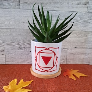 May include: White ceramic planter with a red and white symbol, representing the root chakra, and a green succulent plant inside. The planter is sitting on a wooden coaster on a brown burlap surface.