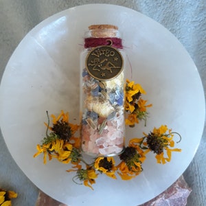 May include: A glass bottle filled with dried herbs, crystals, and salt. The bottle has a cork stopper and a metal charm with the word "Virgo" on it. The bottle is surrounded by dried yellow flowers.
