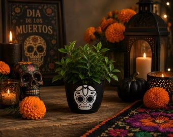 May include: A still life arrangement featuring Day of the Dead decor. A black pot with a sugar skull design holds a green plant. Other items include candles, a skull, orange flowers, and a framed sign that reads "Dia de los Muertos."