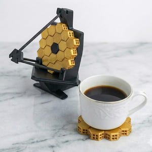 Puede incluir: Un modelo negro y dorado del telescopio espacial James Webb, con una taza de café blanca y un posavasos a juego. El modelo del telescopio presenta un espejo de panal dorado y una estructura de soporte negra. La taza está llena de café oscuro.