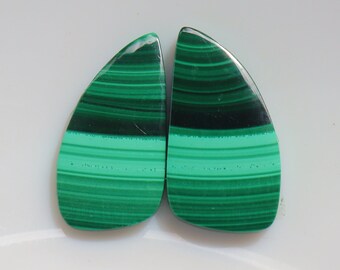 May include: Two green and black malachite cabochons with a striped pattern. The cabochons are teardrop shaped and have a smooth, polished surface.