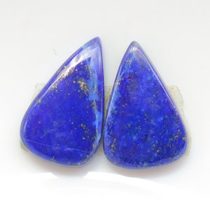 Könnte beinhalten: Zwei tropfenförmige Lapislazuli-Cabochons mit goldenen Flocken. Die Steine sind tiefblau und haben eine glatte, polierte Oberfläche.