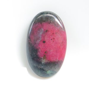 May include: An oval gemstone with a polished finish. The stone displays a bright pink central area, fading into dark grey and black tones at the edges. There are also hints of green. The gemstone is set against a white background.