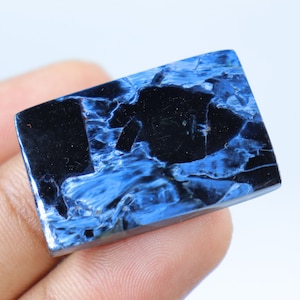 May include: A rectangular gemstone with a polished surface. The stone features a striking pattern of deep blue and black swirls and veins. The gemstone is held in a hand against a white background.