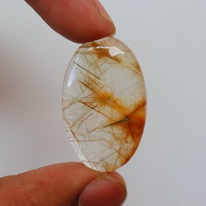 May include: A clear oval-shaped gemstone with a golden-brown rutilated quartz inclusion. The gemstone is held between two fingers.