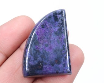 Op de afbeelding: Een paarse en zwarte cabochon edelsteen met een afgeronde rand. De edelsteen wordt in een hand gehouden.