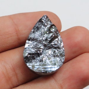 May include: A pearlescent silver teardrop-shaped gemstone with a metallic sheen. The gemstone has a rough, textured surface.