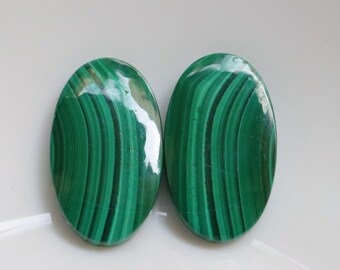 May include: Two oval-shaped malachite stones with a polished, glossy finish. The stones display a vibrant green color with dark green, banded patterns. The gemstones are set against a white background.