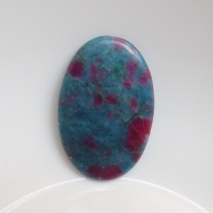 Puede incluir: Piedra preciosa ovalada en cabujón con un patrón moteado azul y rosa. La piedra tiene una superficie lisa y pulida.