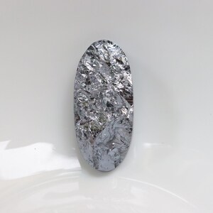 May include: A silvery gray oval-shaped piece of silicon. The silicon has a rough, textured surface.