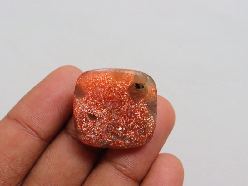 Pu&ograve; includere: Un cabochon di pietra di sole arancione di forma quadrata con una finitura scintillante e iridescente.