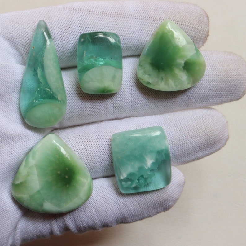 May include: Five green and white cabochon gemstones of various shapes and sizes. The gemstones are arranged on a white surface.