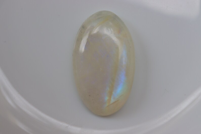 K&ouml;nnte beinhalten: Ein wei&szlig;er ovaler Mondstein-Cabochon mit schillernden blauen und violetten Blitzen.