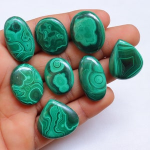 Puede incluir: Una colecci&oacute;n de ocho cabujones de malaquita verde, cada uno con un patr&oacute;n &uacute;nico de remolino. Los cabujones son lisos y pulidos, y est&aacute;n dispuestos en una mano.