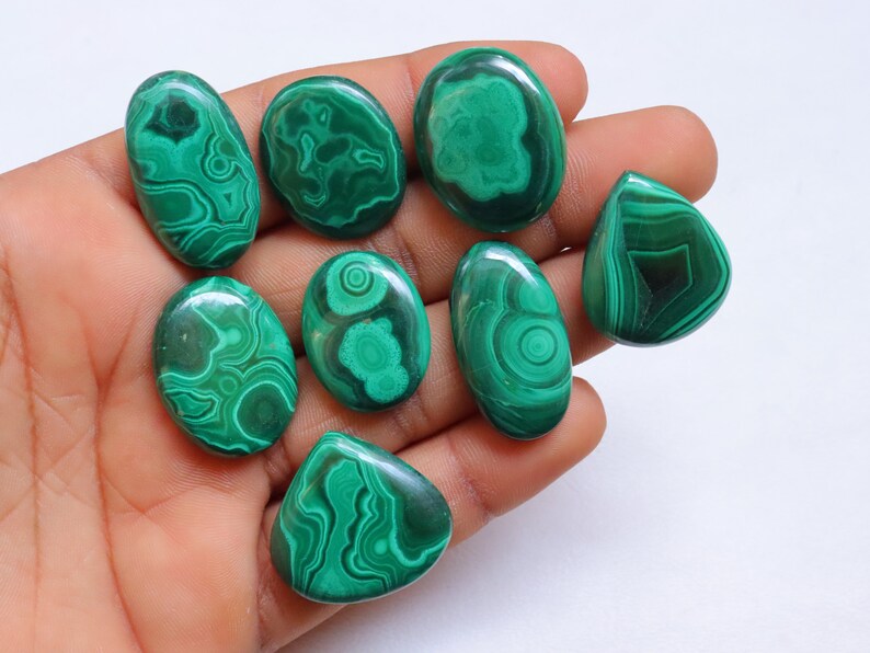 Puede incluir: Una colecci&oacute;n de ocho cabujones de malaquita verde, cada uno con un patr&oacute;n &uacute;nico de remolino. Los cabujones son lisos y pulidos, y est&aacute;n dispuestos en una mano.