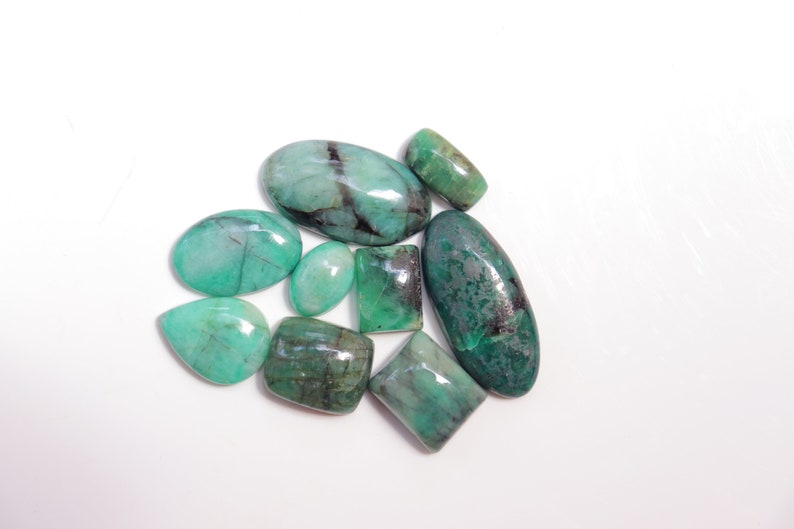 May include: A collection of eight green emerald gemstones of various shapes and sizes. The gemstones are cut and polished, and they have a translucent appearance.