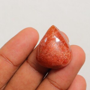 Pu&ograve; includere: Un cabochon di pietra di sole rossa a forma di lacrima con una superficie lucida e lucidata. La pietra ha un aspetto scintillante e scintillante.