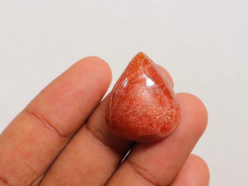 Pu&ograve; includere: Un cabochon di pietra di sole rossa a forma di lacrima con una superficie lucida e lucidata. La pietra ha un aspetto scintillante e scintillante.