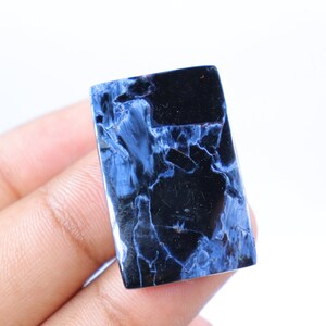 May include: A rectangular gemstone with a polished surface. The stone features a striking pattern of deep blue and black veining, creating a marbled effect. The gemstone is held in a hand against a white background.