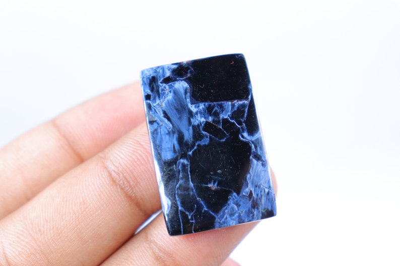 May include: A rectangular gemstone with a polished surface. The stone features a striking pattern of deep blue and black veining, creating a marbled effect. The gemstone is held in a hand against a white background.
